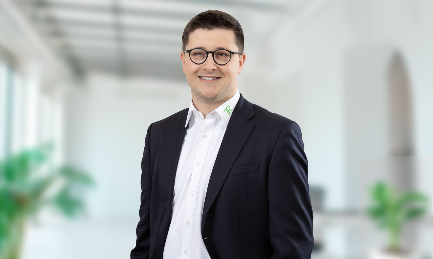 Porträtfoto eines jungen Mannes - Adrian Schulte - in einem modernen, hellen Büro. Er trägt einen dunklen Anzug mit weißem Hemd und eine dunkle Brille. Er lächelt freundlich in die Kamera.