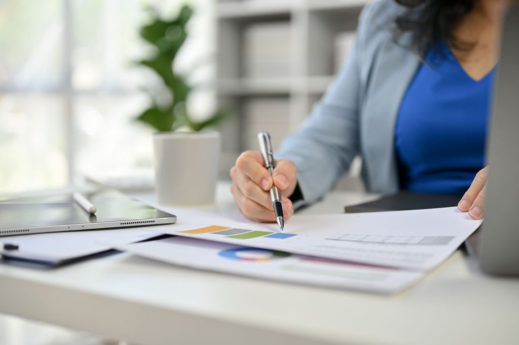 Eine Frau am Arbeitsplatz, sie hält ein Blatt Papier mit farbigen Diagrammen in der Hand und zeigt mit einem Kugelschreiber darauf. Im Hintergrund ist ein Computerbildschirm und eine grüne Pflanze zu sehen. Die Frau trägt ein blaues Oberteil und einen grauen Blazer.
