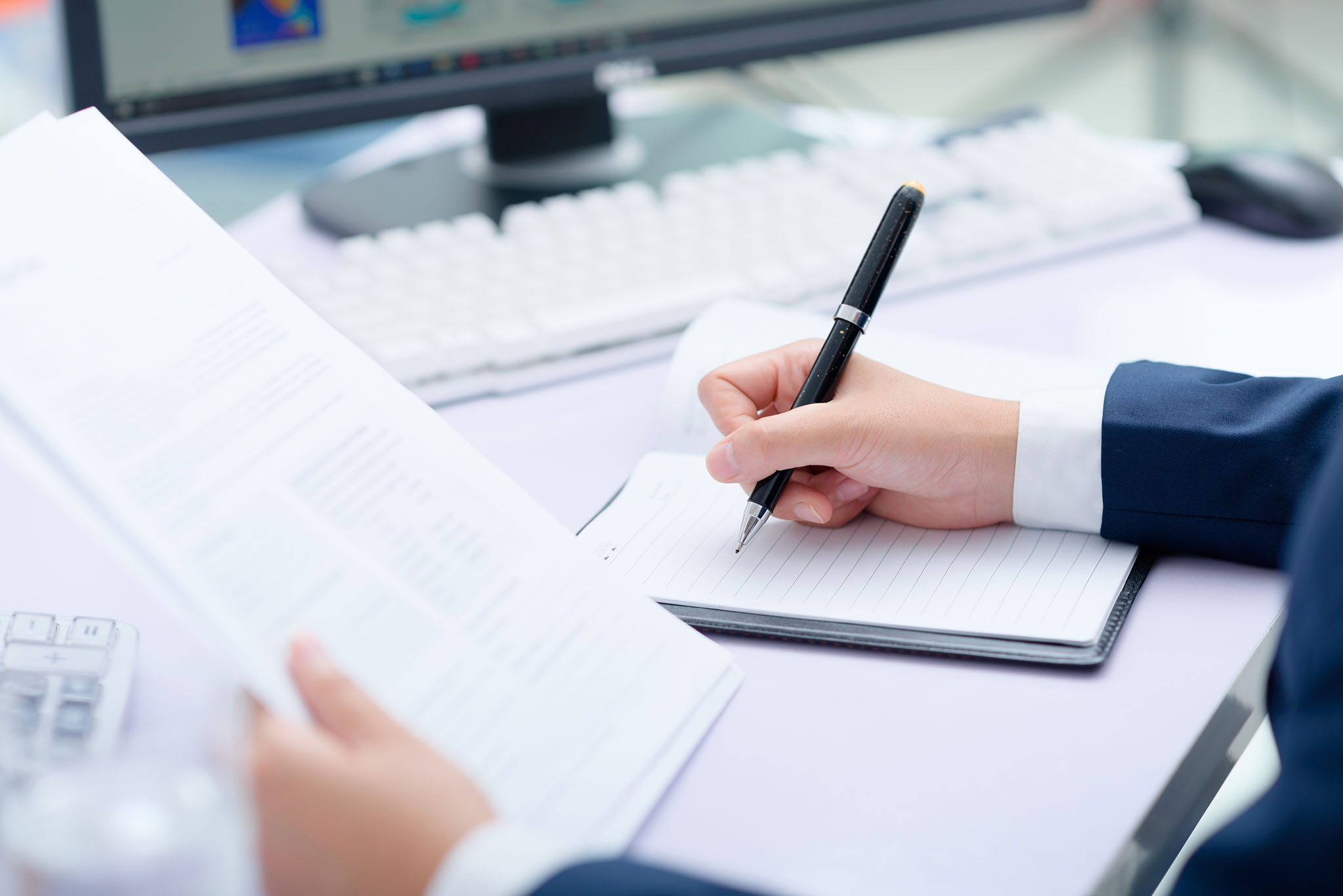 Hand mit Kugelschreiber auf einem Notizbuch. Im Hintergrund befinden sich eine Tastatur und ein Monitor. Die Person macht sich Notizen. Alles ist in hellem Licht gehalten, die Szene wirkt ordentlich und professionell.