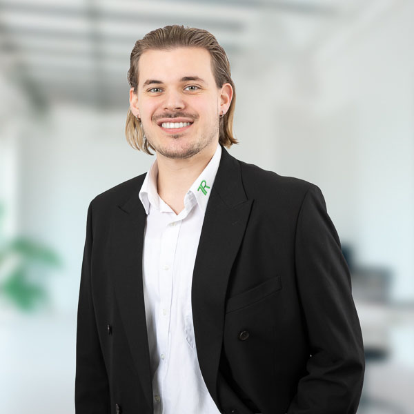 Porträtfoto eines jungen Mannes, Lukas Neubauer. Er hat schulterlanges, nach hinten gekämmtes Haar und trägt einen kurzen Bart. Er lächelt freundlich und schaut direkt in die Kamera. Bekleidet ist er mit einem schwarzen Sakko und einem weißen Hemd, auf dessen Kragen ein grünes Logo zu sehen ist. Der Hintergrund ist unscharf und hell.