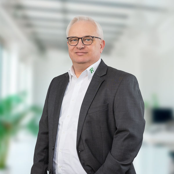 Porträtfoto eines älteren Mannes mit weißem Haar und Brille: Marko Neubauer. Er trägt ein dunkles Sakko über einem weißen Hemd. Auf dem Kragen des Hemdes ist ein grünes Logo zu sehen. Der Mann steht vor einem unscharfen, hellen Hintergrund, der wie ein modernes Büro wirkt.