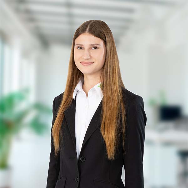 Eine junge Frau mit langem, braunem Haar trägt ein weißes Hemd und einen schwarzen Blazer vor einem unscharfen, hellen Büro-Hintergrund im Porträt.