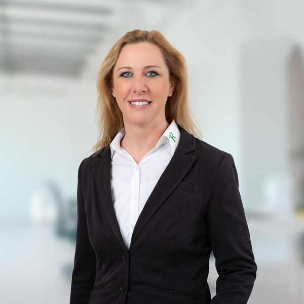 Porträtfoto einer Frau: Melanie Rohrbach. Sie hat schulterlanges, blondes Haar und lächelt freundlich in die Kamera. Auf dem Kragen der Bluse ist ein grünes Logo zu sehen. Sie trägt einen schwarzen Blazer über einer weißen Bluse. Der Hintergrund ist unscharf und hell, ein modernes Büro.