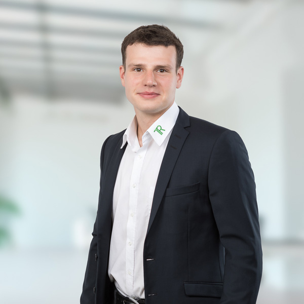 Ein junger Mann in einem Anzug steht in einem modernen Büro mit neutralem Hintergrund und lächelt freundlich in die Kamera.