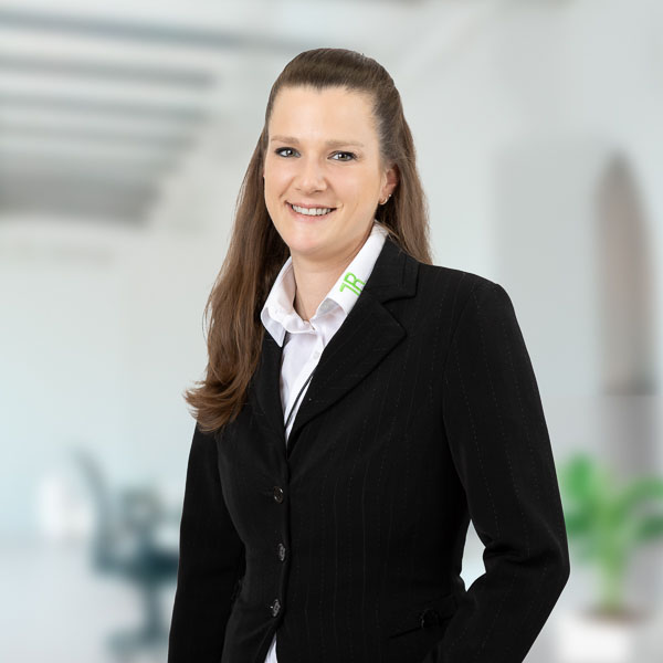 Portätfoto einer Frau in Businesskleidung: Sarah Fischer. Sie hat lange, glatte, braune Haare, die sie nach hinten frisiert trägt. Sie lächelt freundlich und trägt ein schwarzes Jackett über einer weißen Bluse. Auf dem Kragen der Bluse ist ein grünes Logo zu sehen. Der Hintergrund ist unscharf und wirkt wie ein modernes Büro mit hellen Farben und einer Pflanze im Hintergrund.