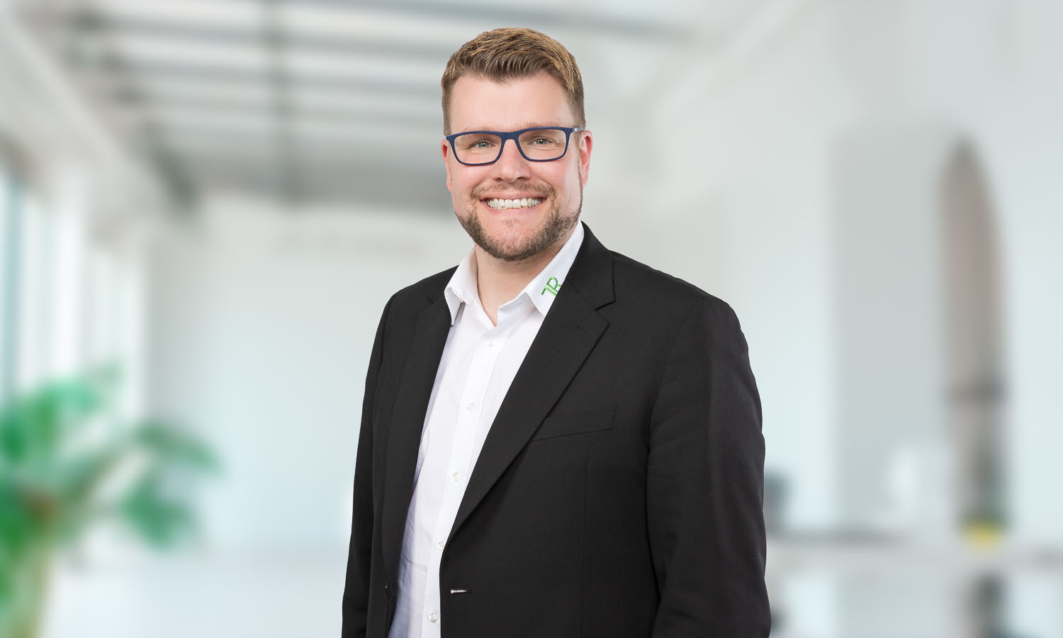 Porträtfoto eines lächelnden Mannes: Tim Rohrbach. Er trägt einen schwarzen Anzug mit weißem Hemd und eine dunkelblaue Brille. Seine Haare sind kurz und blond, er hat einen leichten Bart. Im Hintergrund ist alles unscharf, wohl ein Büro. Auf dem Hemdkragen ist ein grünes Logo "TR" zu sehen.
